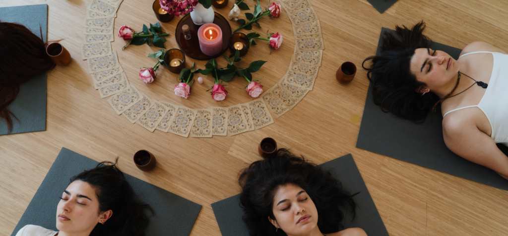 Vrouwen die in een cirkel op yogamatten liggen in een ceremoniële setting met bloemen, kaarsen en kaarten.